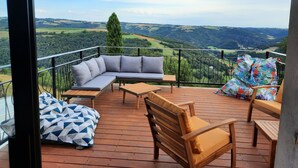 A balcony with a wooden deck, a grey sofa, a wooden chair, and a patterned cushion. - Gîte 12 pers, pétanque court, swimming pool : week from july 11th to 18th free ! (Courris)