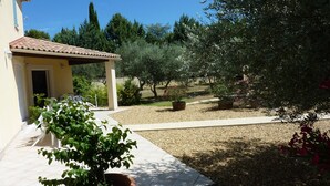 Un espace extérieur aménagé en patio, avec une pergola, des plantes en pot et un chemin de gravier menant à l’entrée d’un bâtiment.