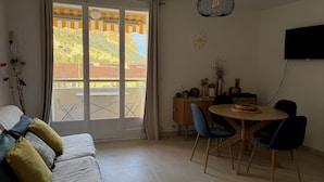 A room with a sofa, a dining table with chairs, a TV mounted on the wall, and a view of a building outside.
