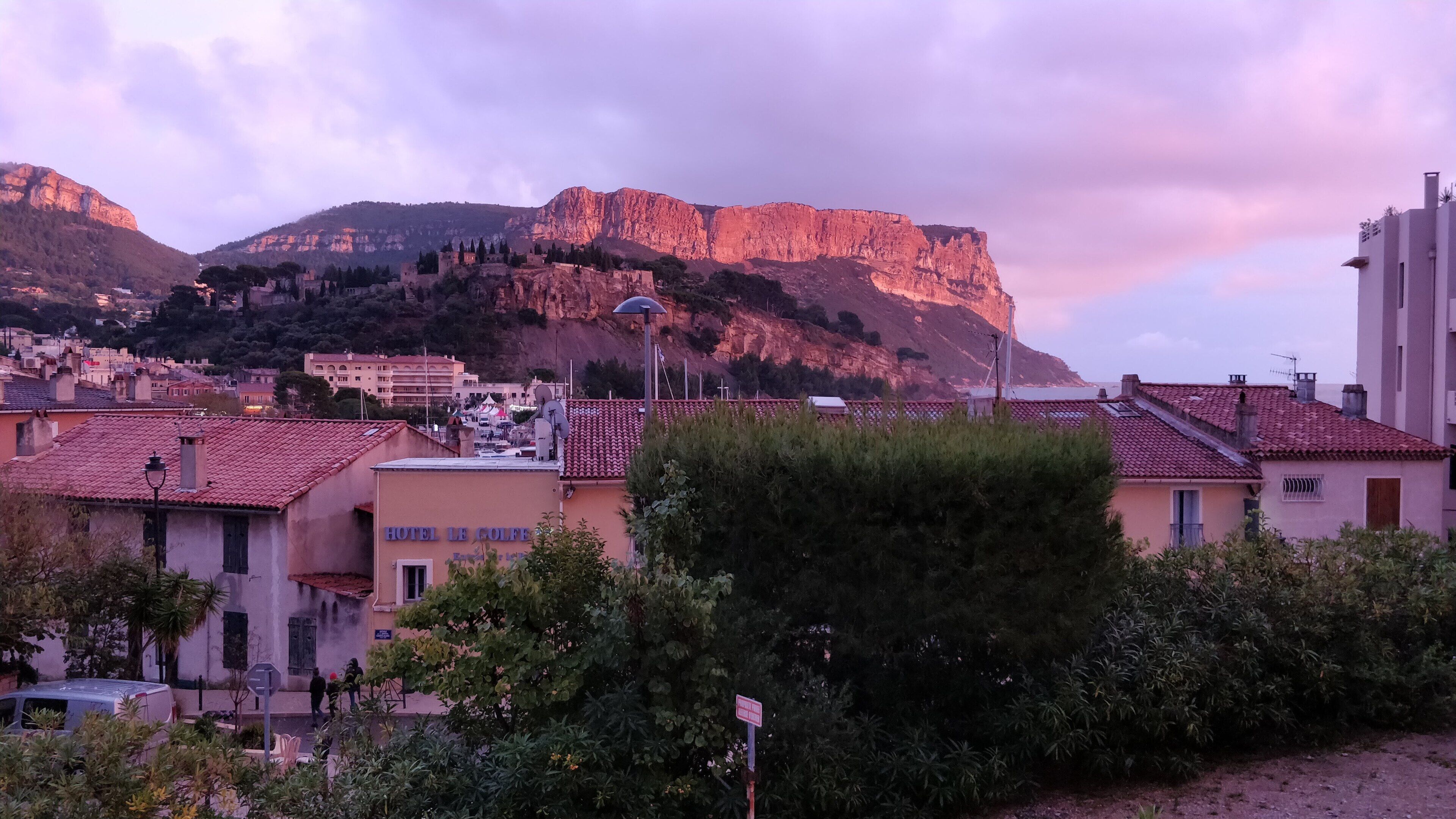 Un hotel amb un rètol que diu "HOTEL LE COLFE", situat en un poble amb teulades vermelles i un paisatge de muntanyes al fons.