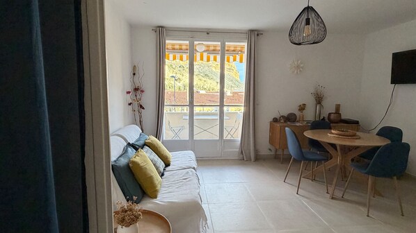 A room with a sofa, dining table, chairs, a hanging light fixture, and a view through a window.