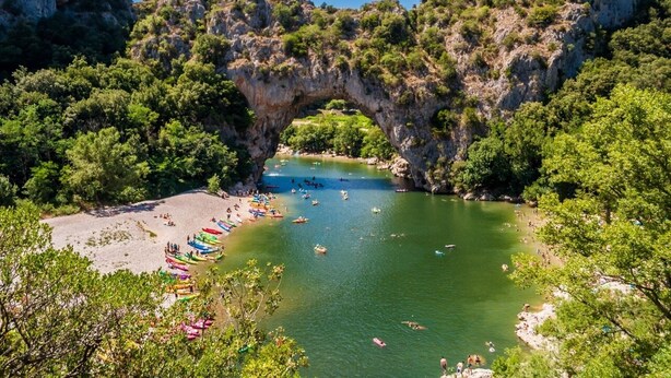 A natural arch formation over a river with boats and people on the shore.