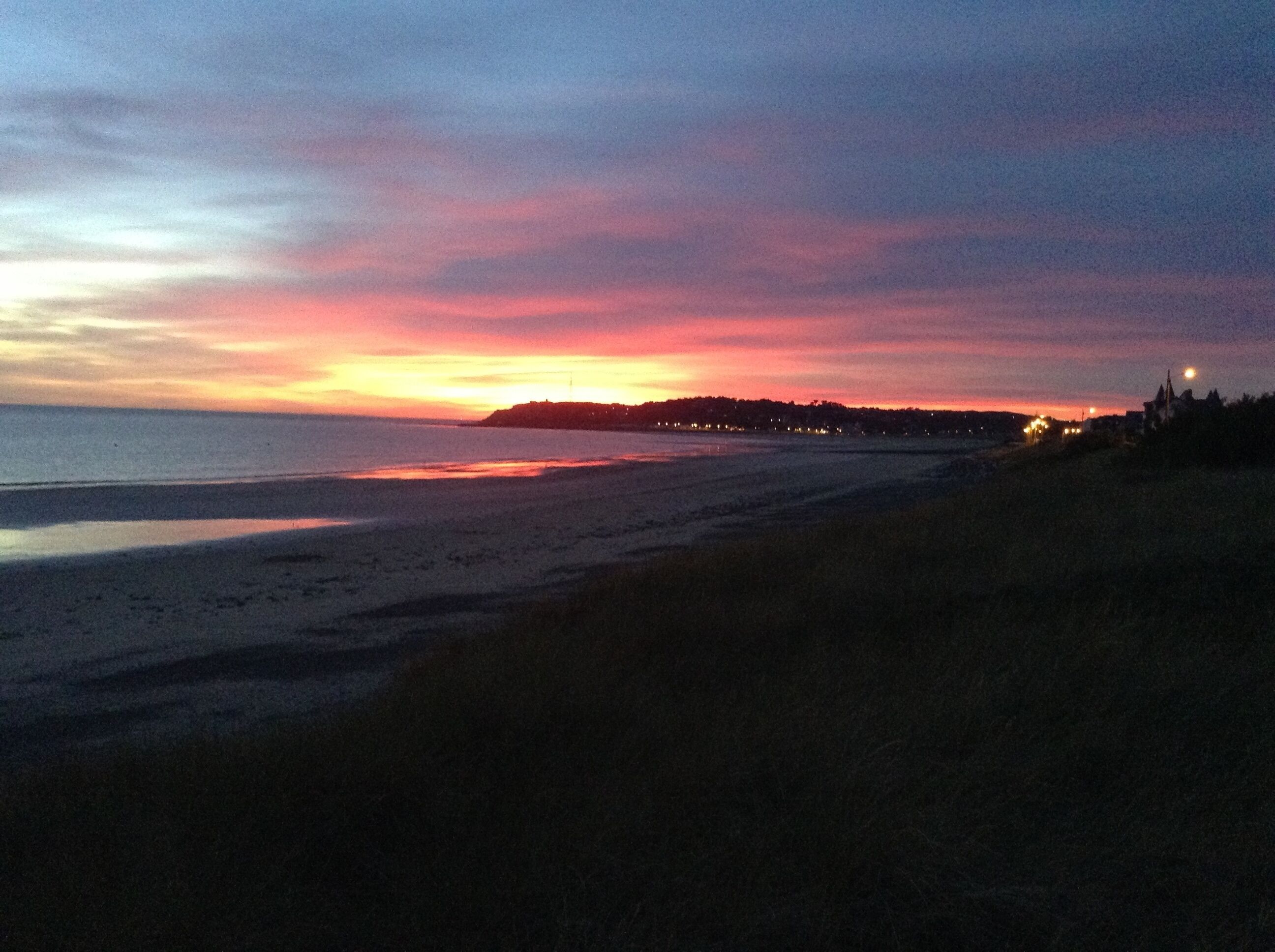 Una playa al atardecer con una costa lejana y algunos edificios.