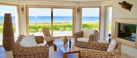 Een kamer met rieten meubels, een glazen salontafel en grote ramen met uitzicht op het strand.