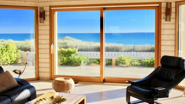 A room with a large sliding glass door leading to a beach view, a black leather chair, a wooden coffee table, and a sofa with a cushion.