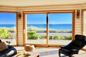 A room with a large sliding glass door leading to a beach view, a black leather chair, a wooden coffee table, and a sofa with a cushion.