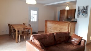 A living room with a brown leather sofa, a wooden dining table with chairs, a kitchen area with wooden cabinets, and a window with a view of the street.