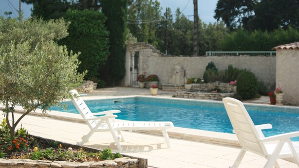 A swimming pool with two white lounge chairs, surrounded by greenery and a stone wall. - House located in the countryside (Mollégès)