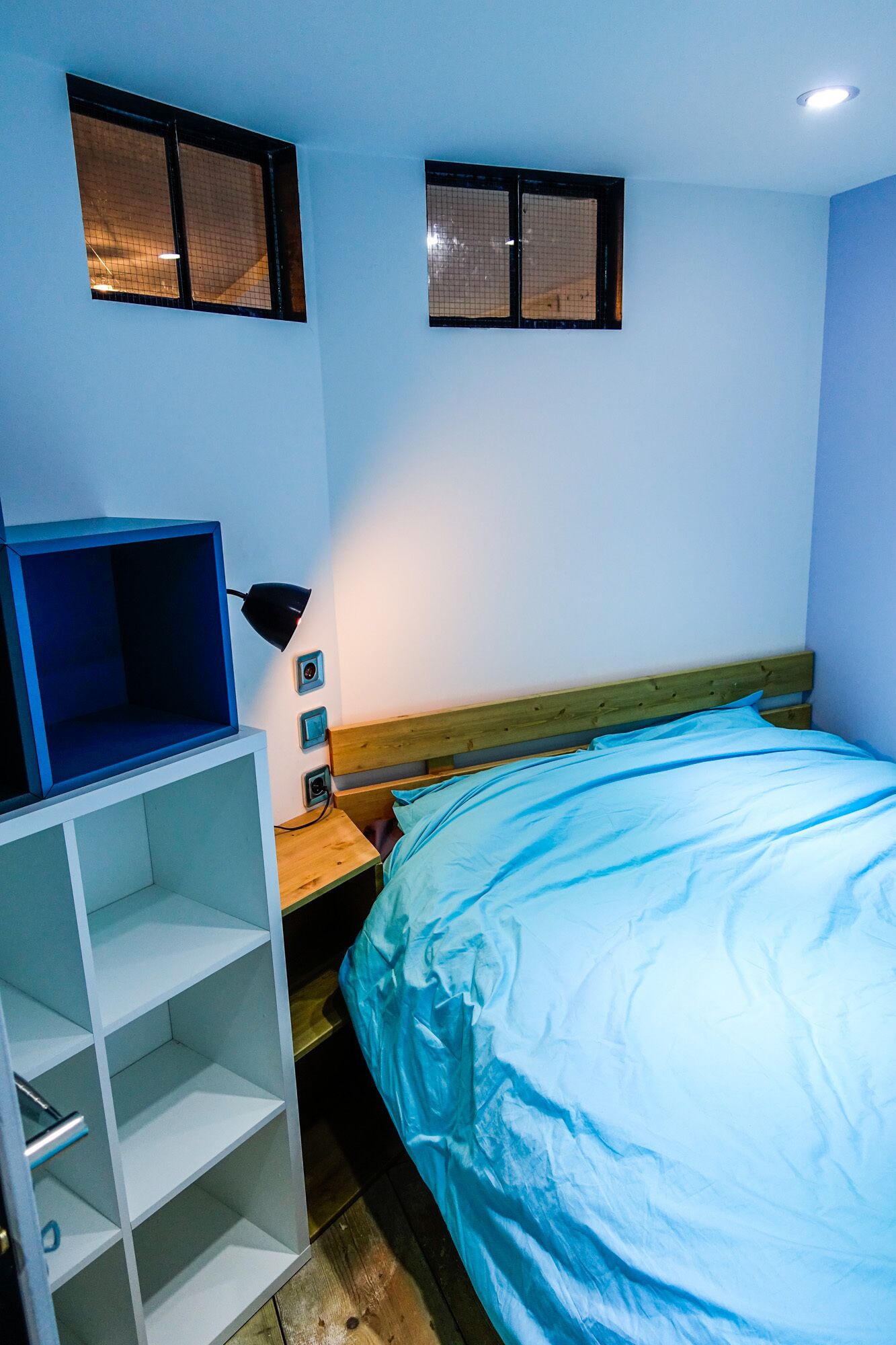 A bedroom with a bed, a desk with a chair, a shelf unit, and two windows.