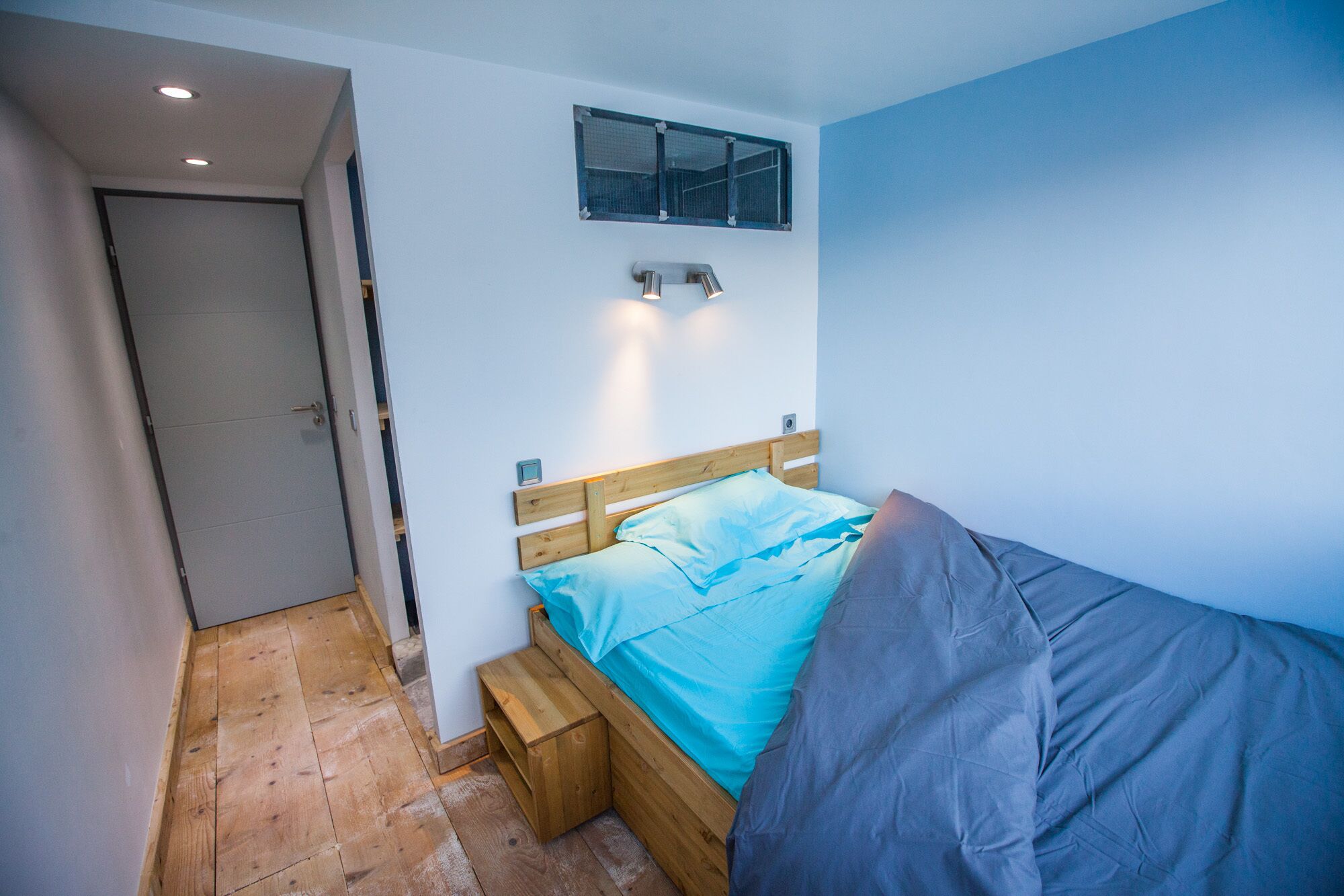 Ein kleines Zimmer mit einem Einzelbett, einem hölzernen Kopfteil, einem kleinen Fenster und einer Tür.