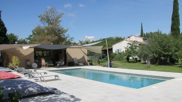 Une piscine rectangulaire, agrémentée de chaises longues et de parasols, entourée d’un jardin et d’une maison dont le toit est recouvert de tuiles.