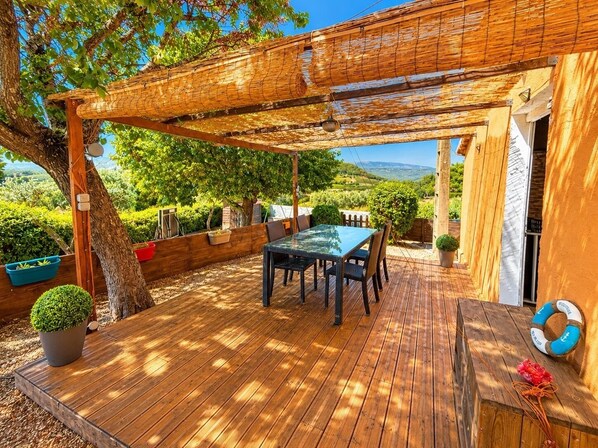 A wooden deck with a table and chairs, surrounded by greenery and a view of the countryside.