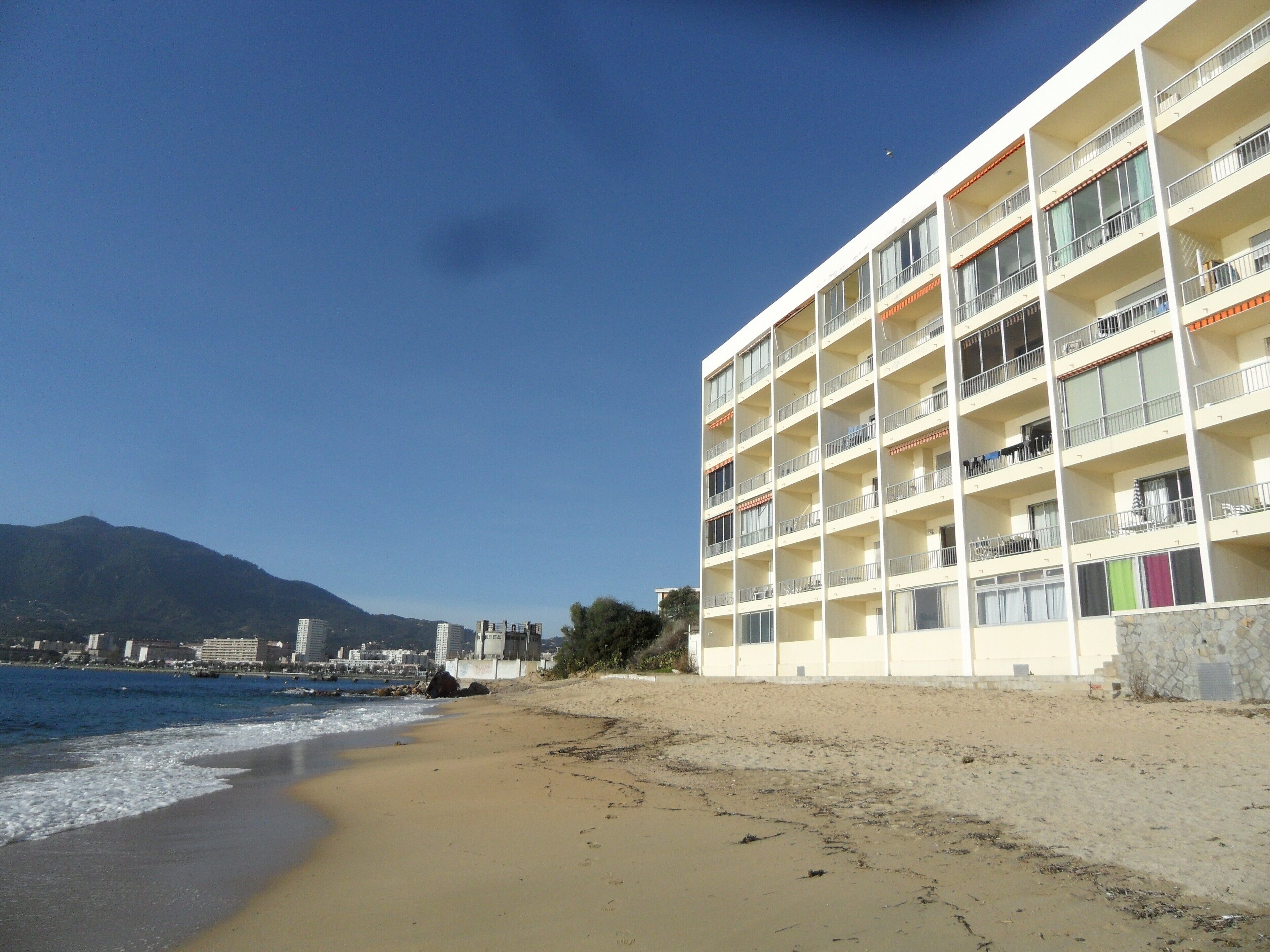 A beachfront hotel with multiple balconies, a sandy beach, and a mountain in the background.
