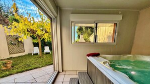 Een hottub met een houten bank, een raam met uitzicht op een tuin met stoelen, en een betegeld terras.
