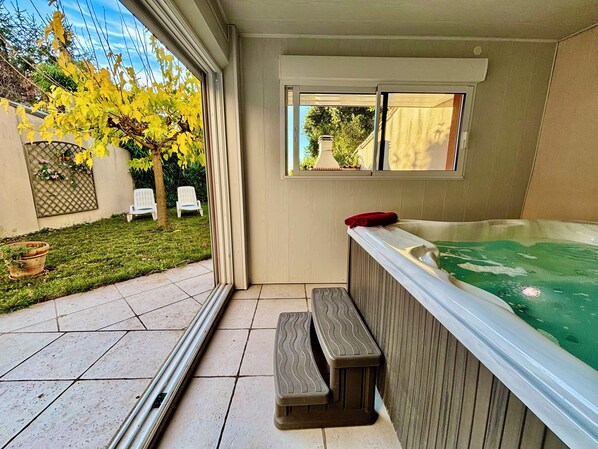 A hot tub with a wooden bench, a window overlooking a garden with chairs, and a tiled patio.