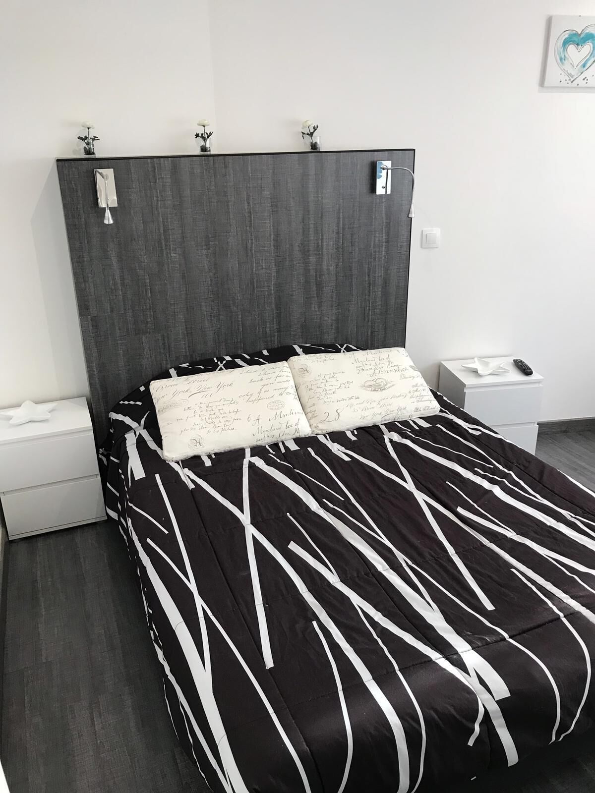 Une chambre à coucher moderne avec une tête de lit sombre, une couvre-lit à rayures noires et blanches et une table de chevet dotée d’une télécommande.
