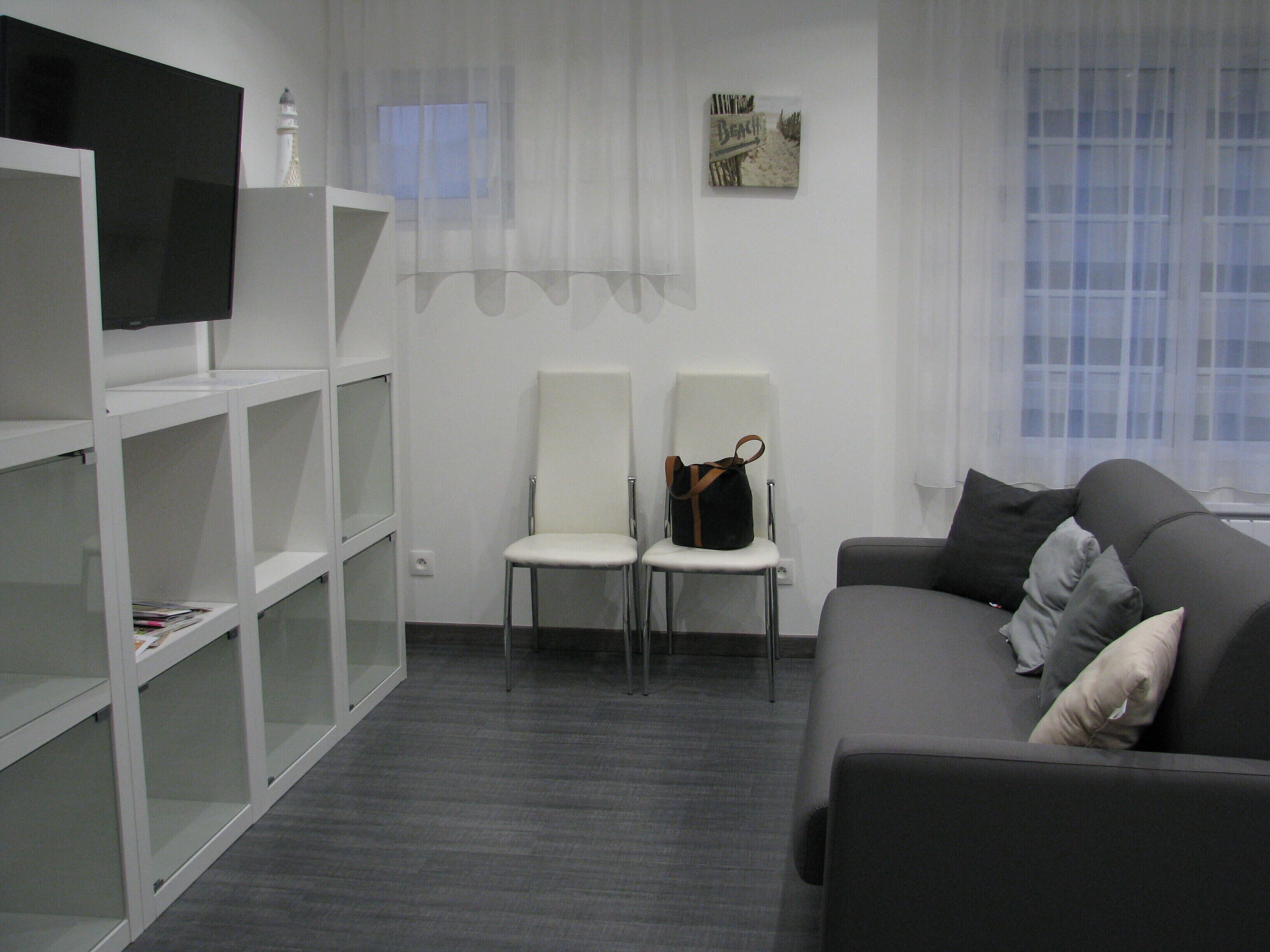 A modern living room with a grey sofa, two white chairs, a TV on a white shelf, and a beach-themed artwork on the wall.