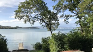 A stone patio overlooking a calm lake with a wooden dock, surrounded by lush greenery and distant hills. - T4 design in lush greenery, sea view and swimming pool, 10 minutes from the beaches.  (LES TROIS ILETS)