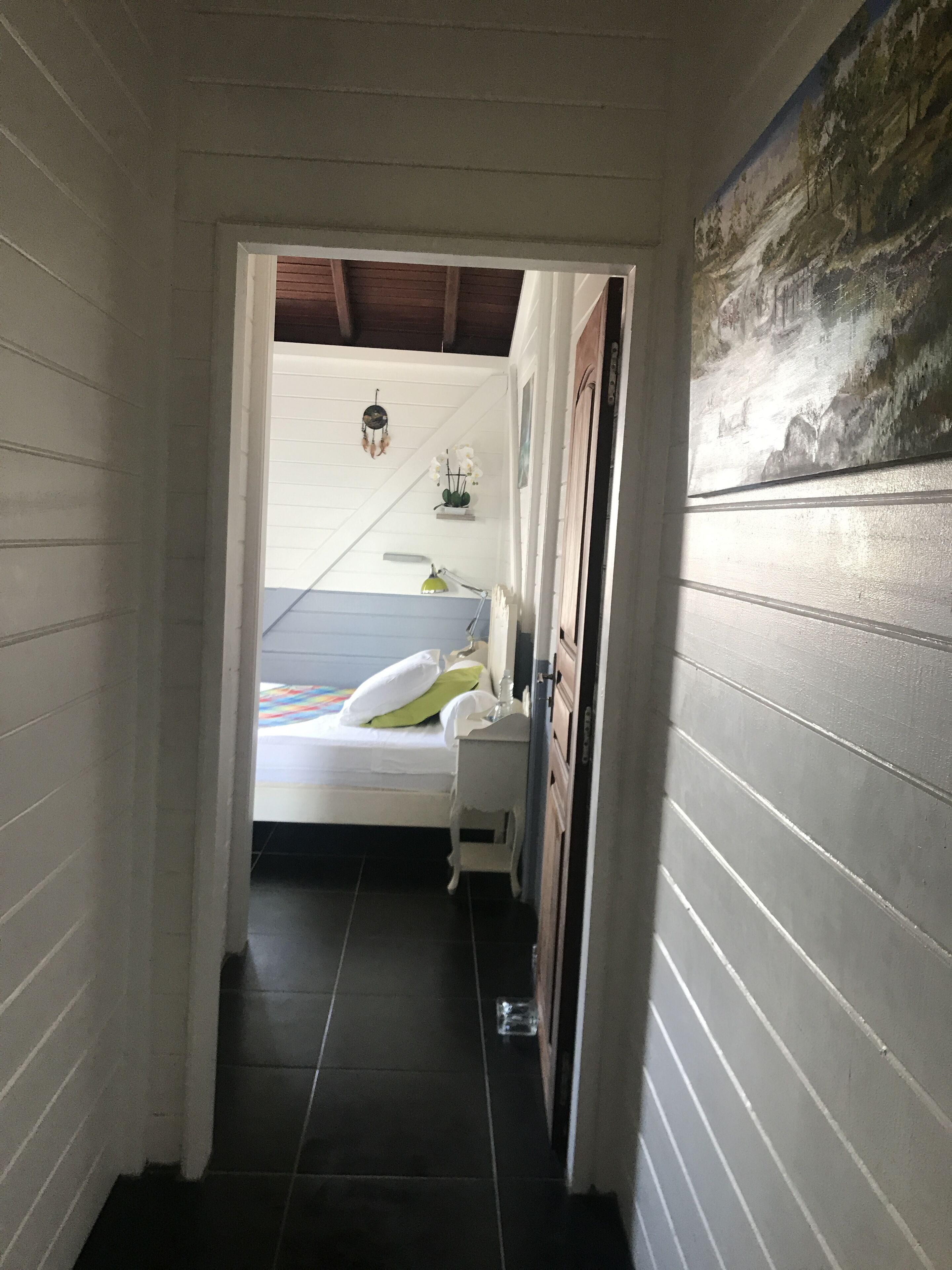 A narrow hallway leading to a bedroom with a bed, bedside table, and a wall-mounted shelf with decorative items.