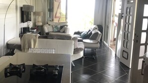 A spacious living area with a kitchen counter, a dining table, and a sofa. There is a large window providing natural light and a view of the outdoors. - T4 design in lush greenery, sea view and swimming pool, 10 minutes from the beaches.  (LES TROIS ILETS)