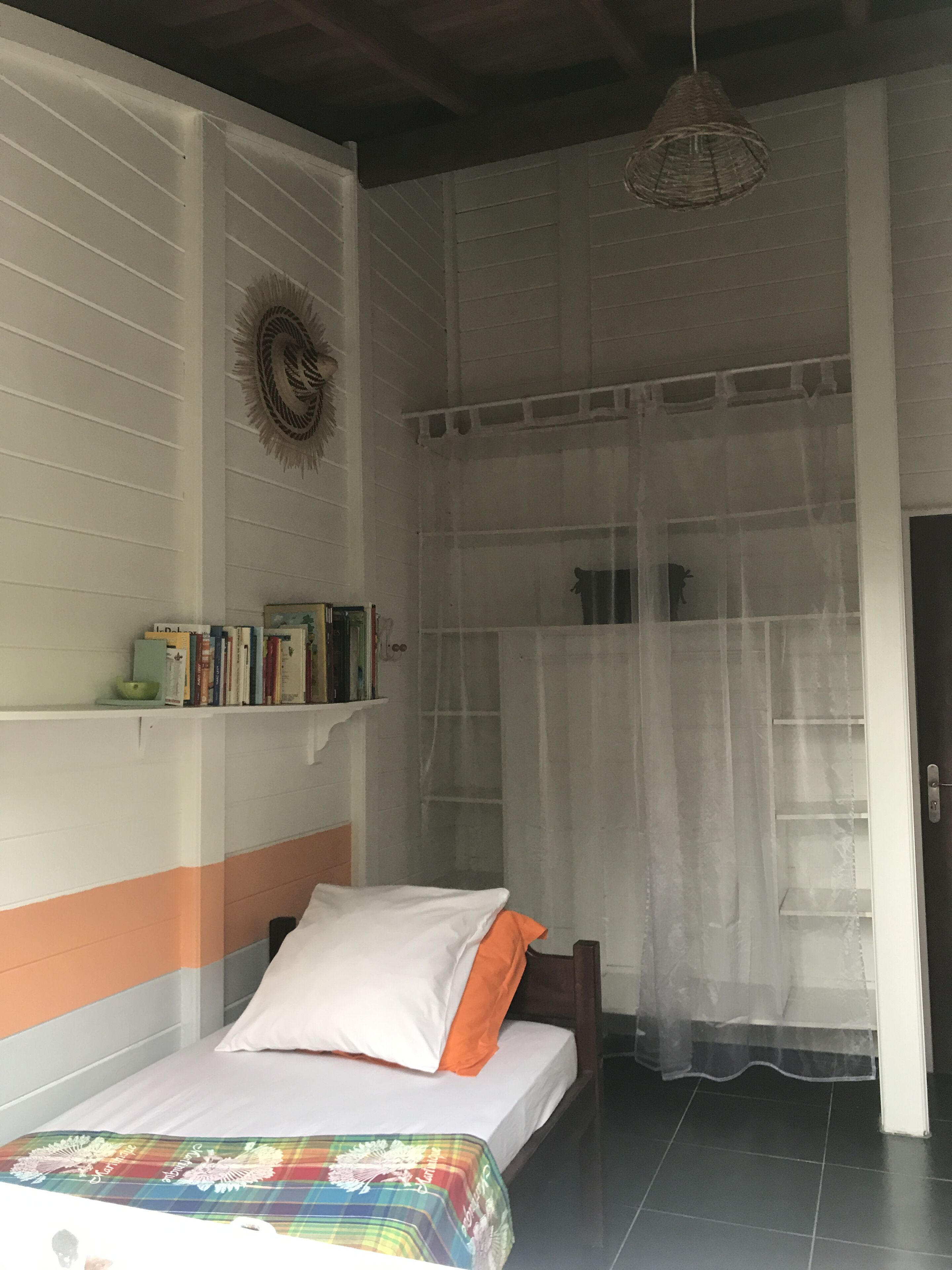 Une pièce comprenant un lit, une étagère remplie de livres, un élément décoratif fixé au mur et une fenêtre ornée d’un rideau léger.