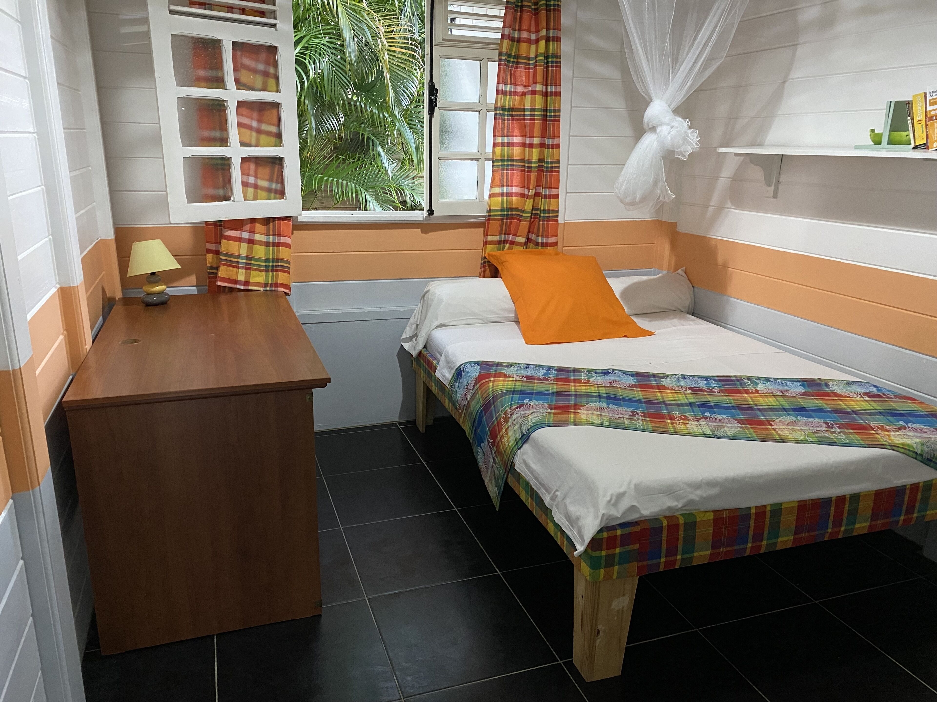 A small, neatly arranged room with a bed, a bedside table, a lamp, a window with curtains, and a shelf with books.