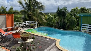 A pool area with lounge chairs, a wooden deck, and a view of palm trees and mountains. - T4 design in lush greenery, sea view and swimming pool, 10 minutes from the beaches.  (LES TROIS ILETS)