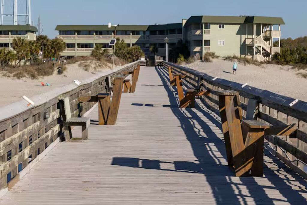 Une promenade en bois menant à une plage, avec une tour de surveillance des sauveteurs et des bâtiments en arrière-plan.