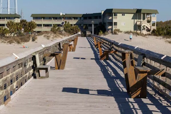 A wooden boardwalk leading to a beach with a lifeguard tower and buildings in the background.