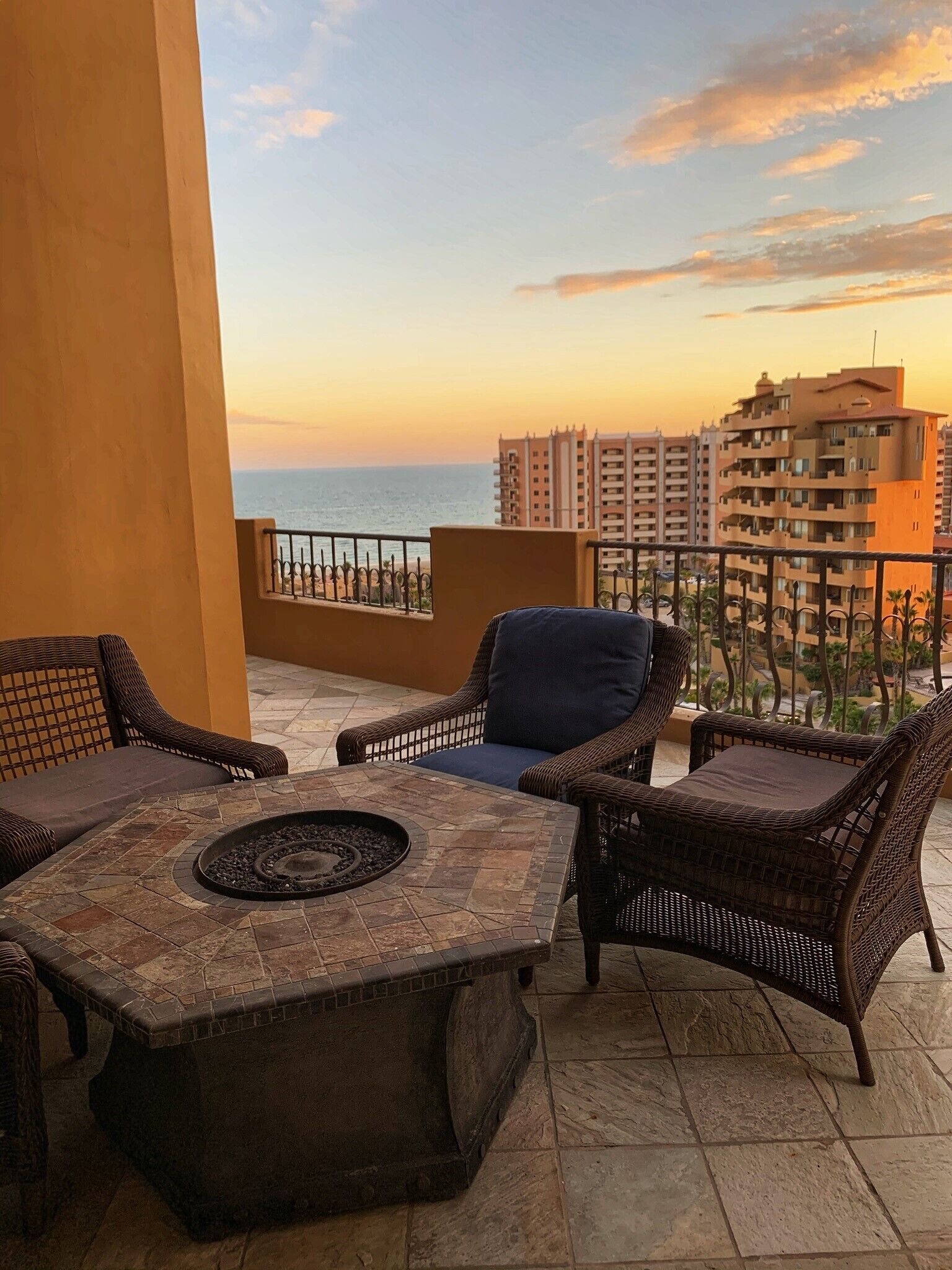 A balcony with wicker furniture, a stone fire pit, and a view of the ocean and buildings at sunset.