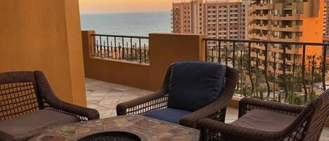 A balcony with wicker furniture, a stone fire pit, and a view of the ocean and buildings at sunset.