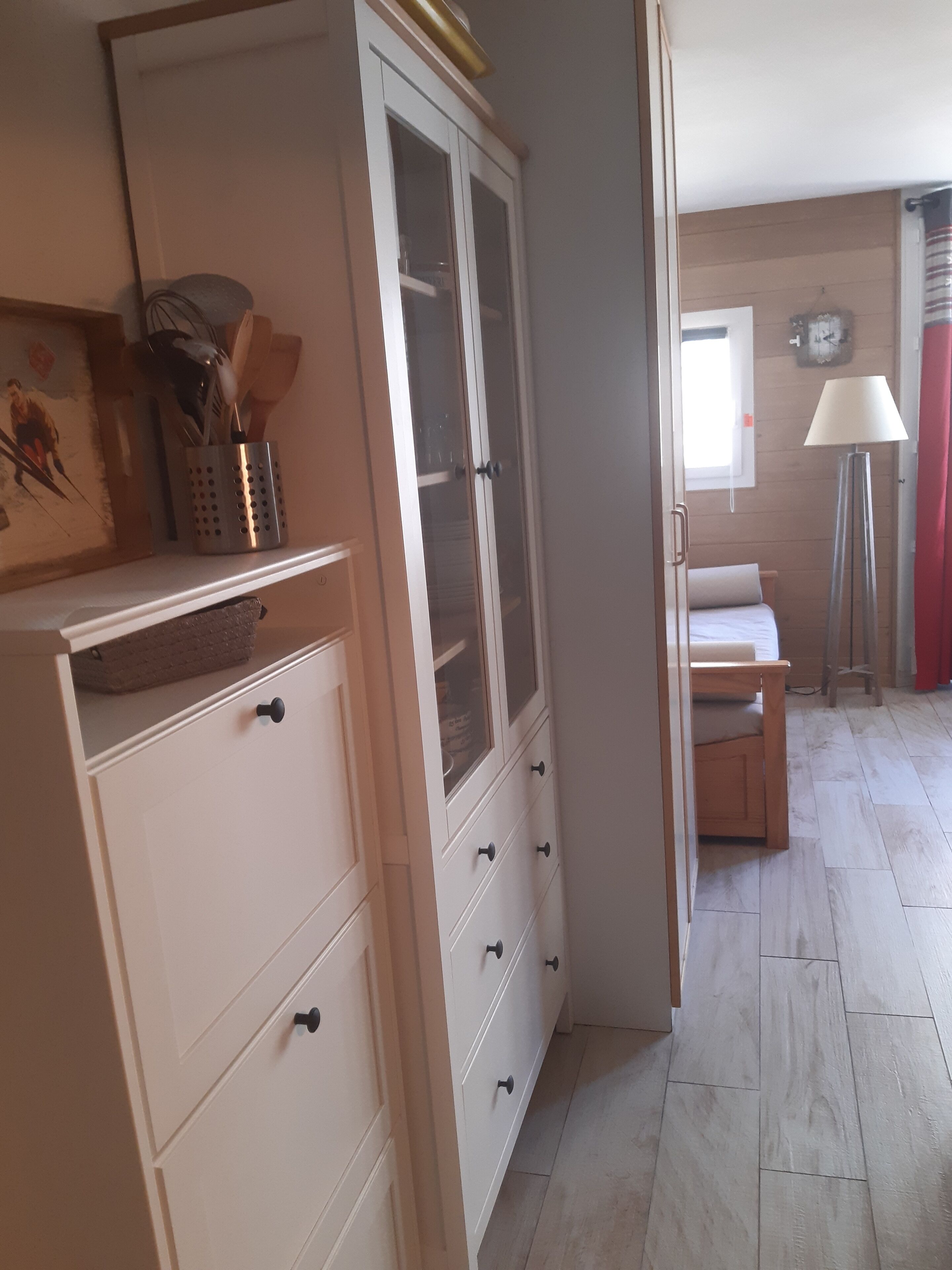 Una habitación con un armario blanco con cajones, una cama de madera, una lámpara de pie y una ventana con cortinas.