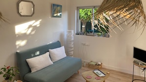 A living room with a sofa, a television, a window with plants, and a hanging plant. - Small house, air conditioning and shaded garden, in a quiet area near the beach (Fleury)