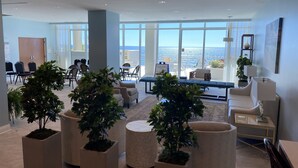 Un hall d’entrée spacieux offrant une vue sur l’océan, avec un espace salon comprenant des tables rondes, des plantes en pot et une grande fenêtre donnant sur la mer.