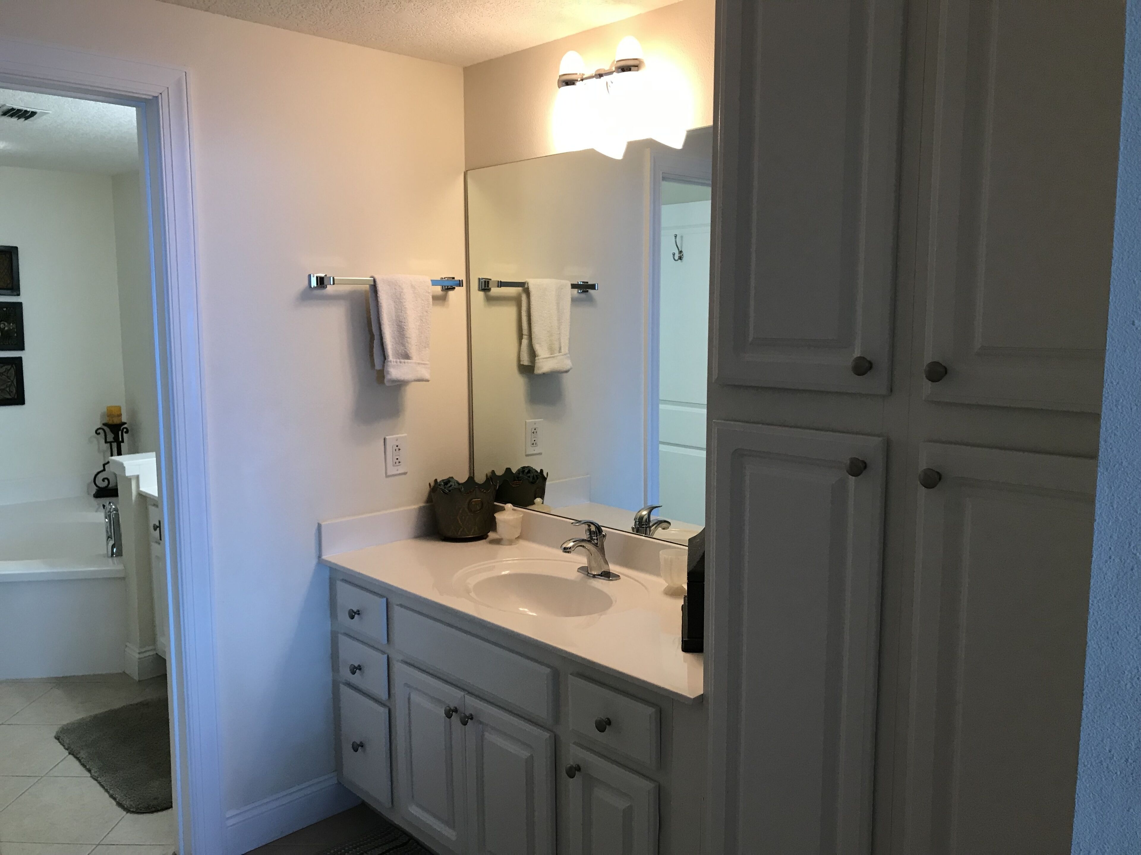 A bathroom with a white sink, mirror, and cabinets. There is a towel rack above the sink and a door with a handle.