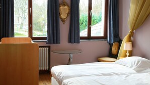 A hotel room with a bed, a wooden chair, a small table, a lamp, and a window with curtains.