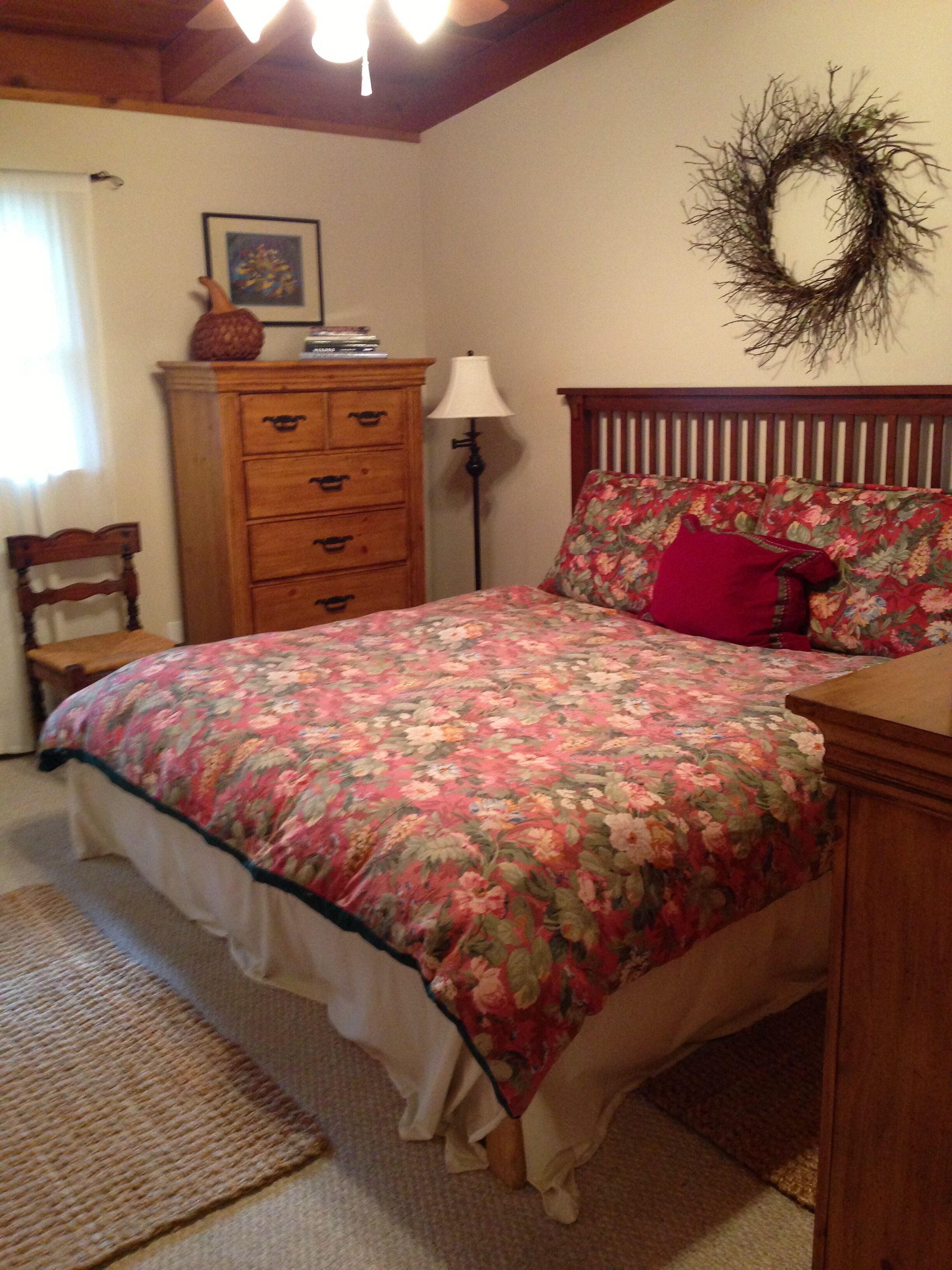 A bedroom with a floral bedspread, a wooden dresser, a chair, a lamp, and a wall wreath.