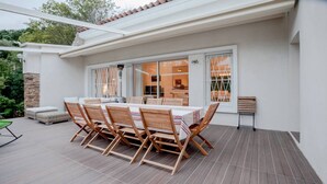 Une terrasse avec une table et des chaises en bois, un pilier en pierre et une vue sur l’intérieur à travers des portes vitrées.