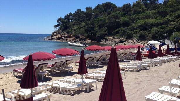 Une plage bordée de rangées de chaises longues blanches et de parasols rouges, un littoral rocheux et un ciel bleu azur.