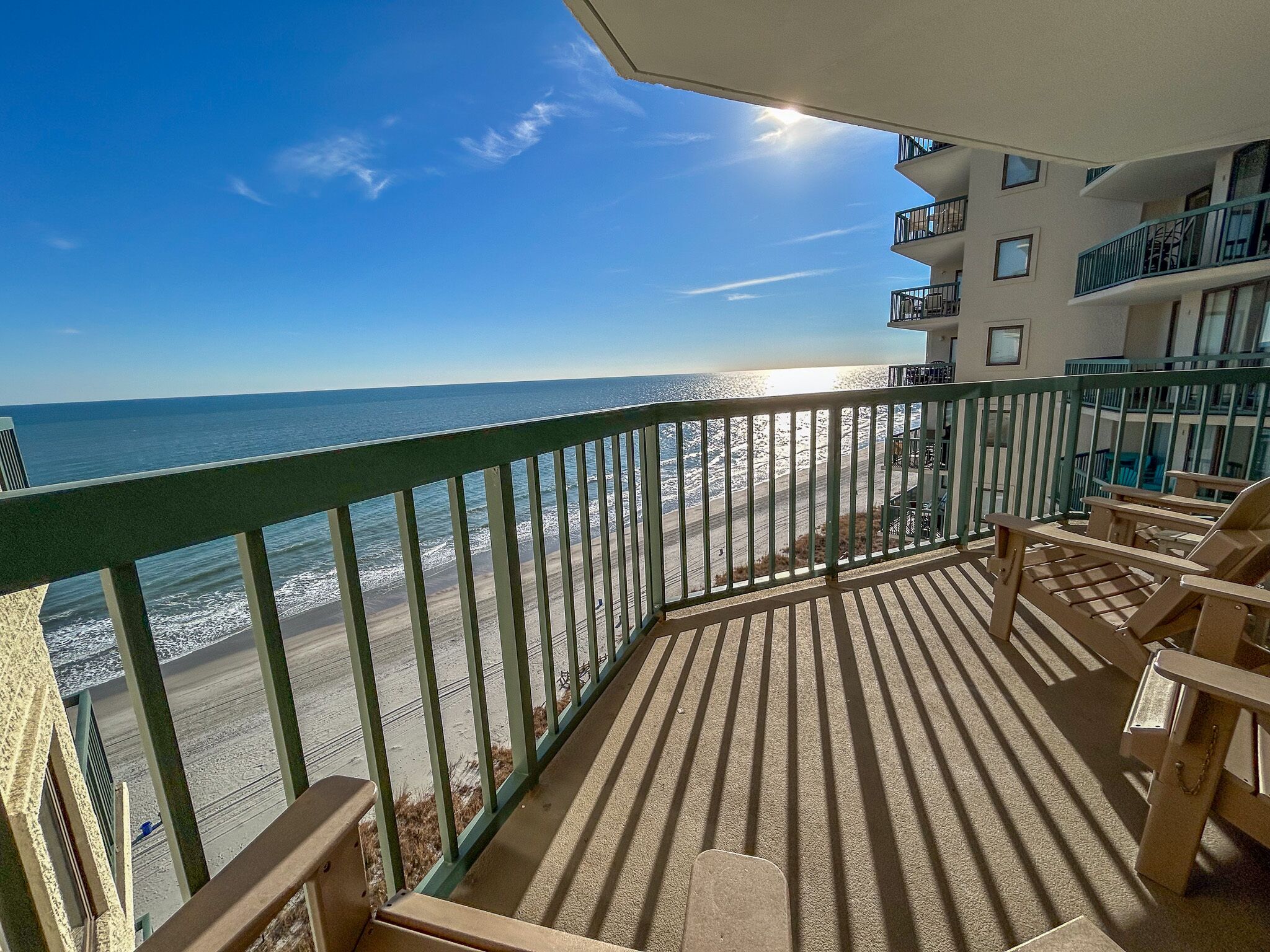 A balcony with a view of the ocean, wooden chairs, and a building with balconies.