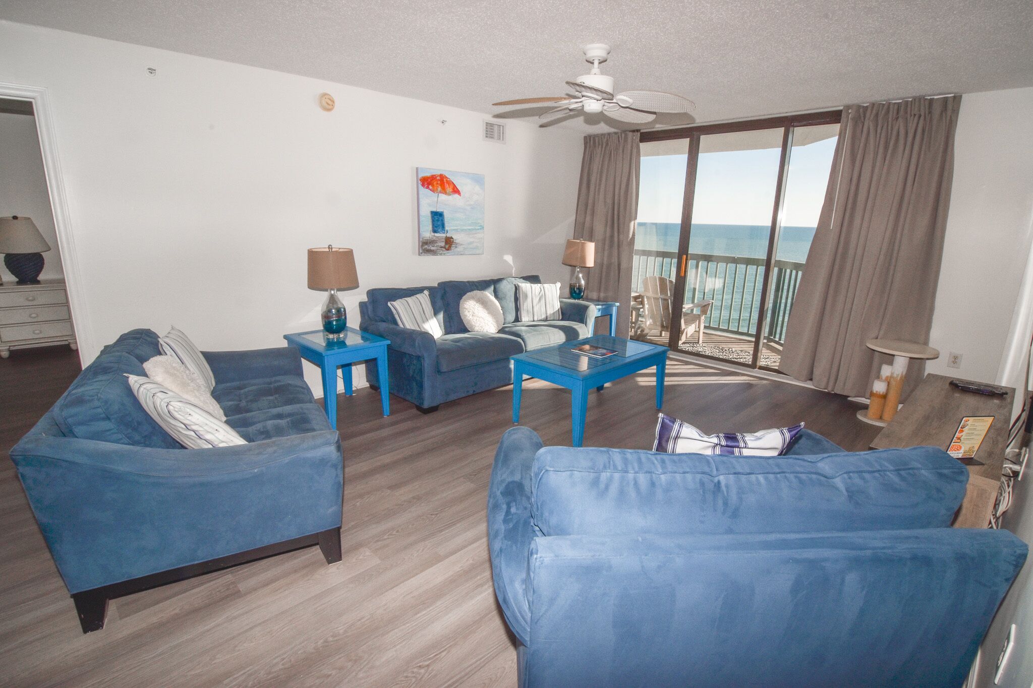 A living room with blue sofas, a coffee table, and a ceiling fan. There is a large window with a view of the ocean.