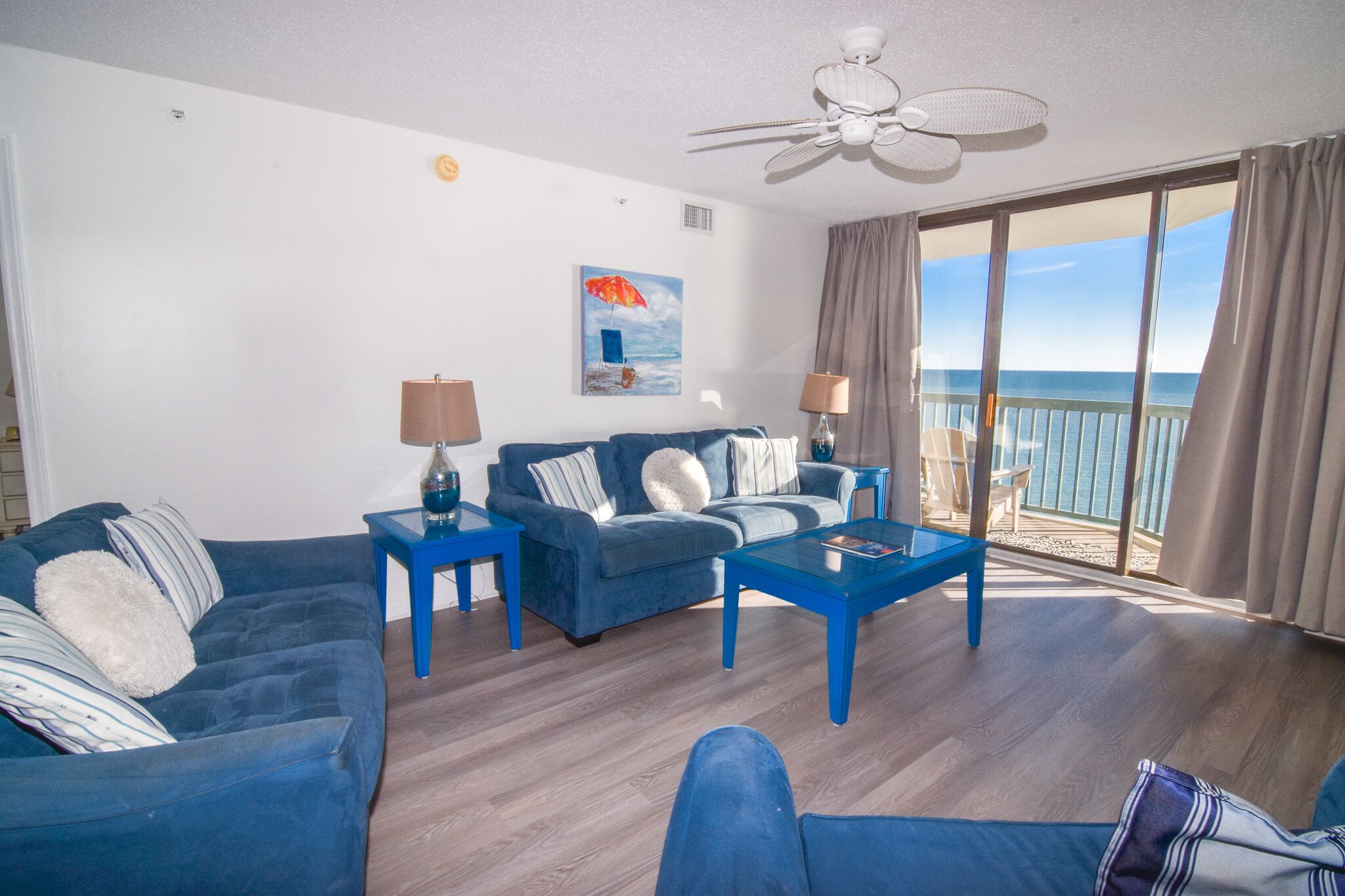 A living room with a blue sofa set, two side tables with lamps, a coffee table, and a large window with a view of the ocean.
