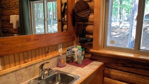 A wooden cabin bathroom with a sink, mirror, and window overlooking a forest. - Sha Bear Cabin, on the YAAK RIVER~Quiet, Romantic Cabin Sleeps up to 4 (Troy)
