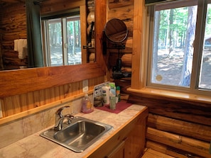 A wooden cabin bathroom with a sink, mirror, and window overlooking a forest.