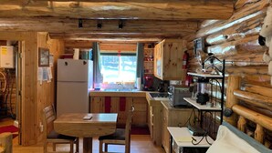 A cozy cabin interior with a wooden dining table, chairs, a kitchen area with a sink, and a window providing a view of the outdoors. - Sha Bear Cabin, on the YAAK RIVER~Quiet, Romantic Cabin Sleeps up to 4 (Troy)