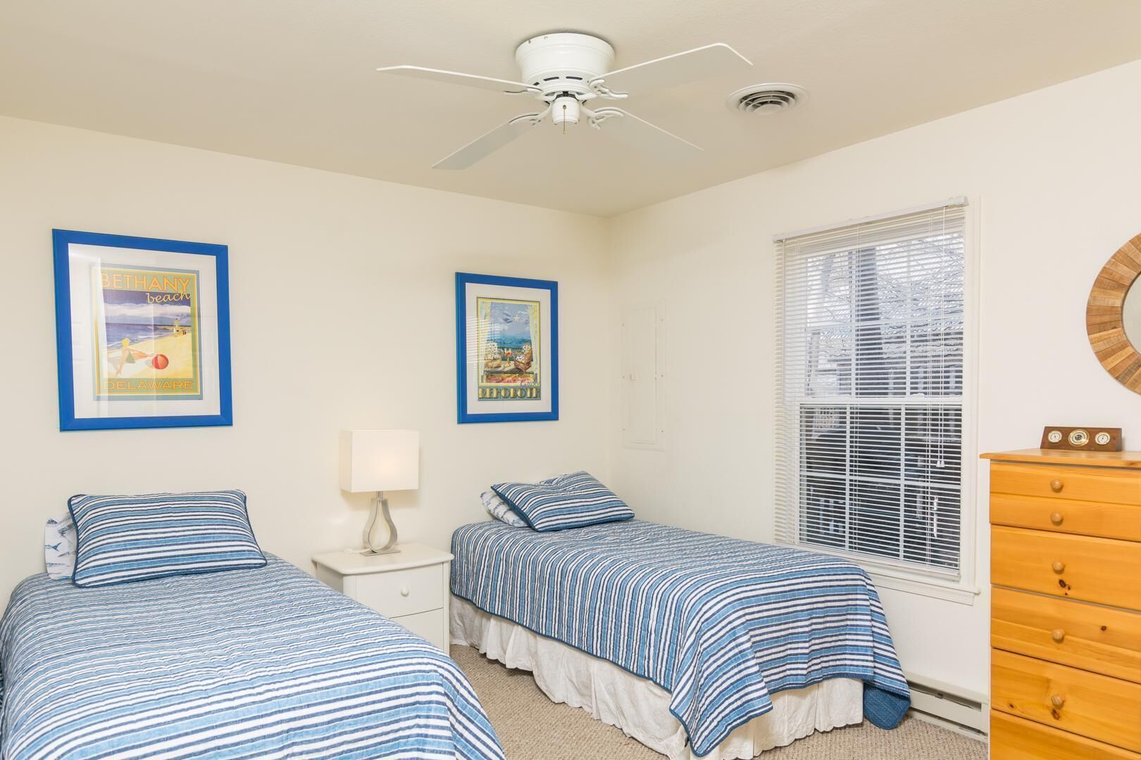 A bedroom with two beds, a ceiling fan, framed pictures on the wall, a window with blinds, and a wooden dresser.