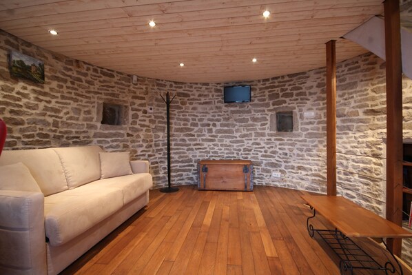 Ein Zimmer mit einer Steinswand, Holzboden, einem weißen Sofa, einem Holztisch und einem an der Wand montierten Fernseher.
