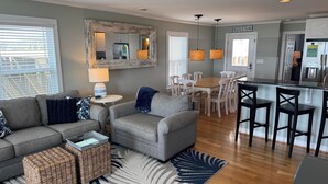 A living room with a grey sofa, a small table with a lamp, a wicker basket, and a dining area with a wooden table and chairs. - Oceanfront Home (Holden Beach)