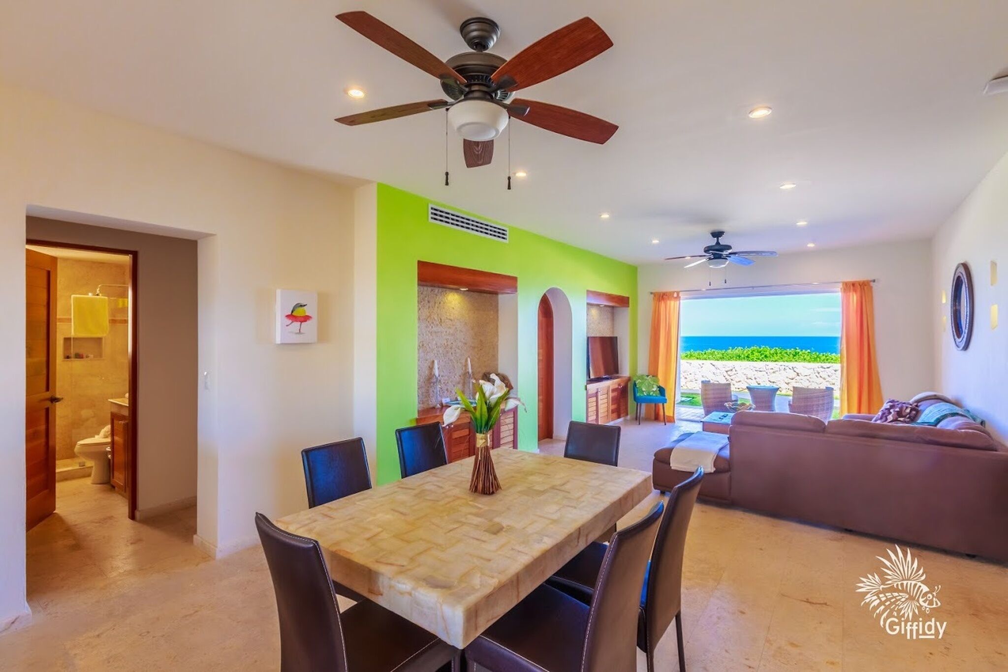 A dining area with a table, chairs, and ceiling fans. There is a view of the beach through the window.