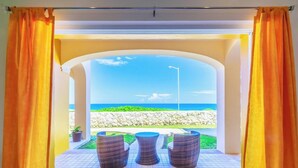 Ein Außenbereich mit drei Korbstühlen und einem Tisch, einer schwarzen Matte und Blick auf das Meer durch ein rundbogenförmiges Fenster mit orangefarbenen Vorhängen.